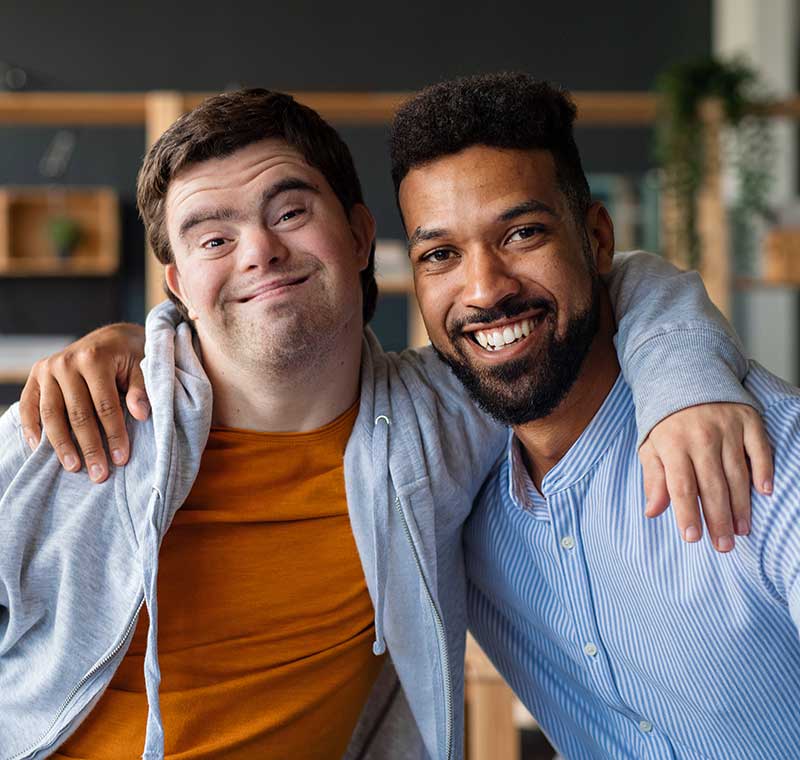 Young man with Down syndrome and his tutor with arms around looking at camera, smiling