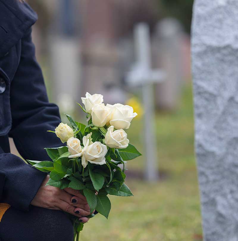 Partial view of woman, her hands holding flowers in front of a grave site.